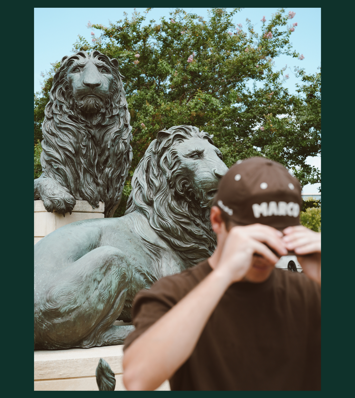 A man wearing a brown t-shirt and a brown baseball cap with 'MARCO' printed on the front stands in front of a bronze lion statue in an outdoor setting with trees in the background.